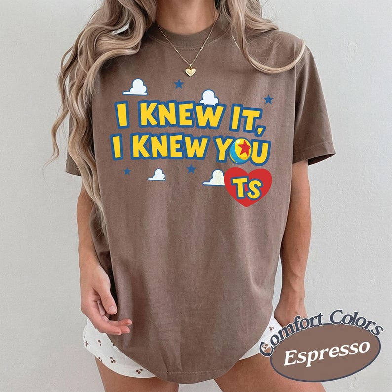 K&ouml;nnte beinhalten: Ein braunes T-Shirt mit kurzen &Auml;rmeln, gelbem Text I KNEW IT, I KNEW YOU, einem roten Herz mit den Buchstaben TS sowie blauen Wolken- und Sternmotiven. Das T-Shirt hat einen Rundhalsausschnitt und eine lockere Passform mit Grafik auf der Vorderseite.