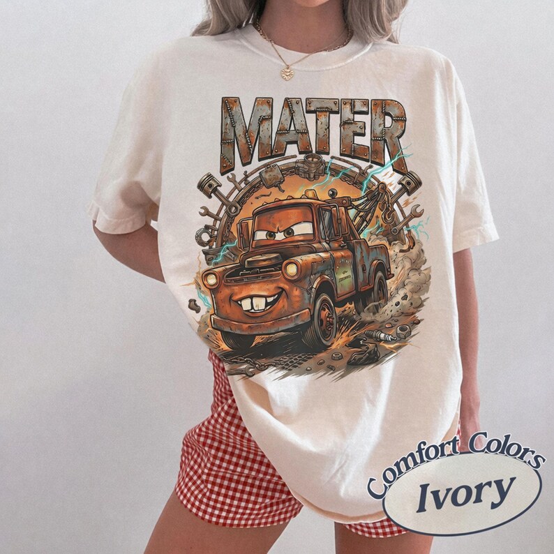 May include: An ivory t-shirt featuring a graphic print of the character Mater from the movie Cars. The design includes the text MATER in a metallic font, a rusty tow truck, mechanical parts, and lightning bolts. The shirt has a crew neckline.