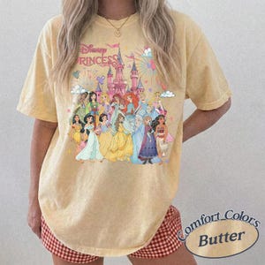 May include: Butter-colored t-shirt featuring a graphic of Disney Princesses in front of a castle. The text "Disney Princess" is above the castle. The shirt is a Comfort Colors brand.