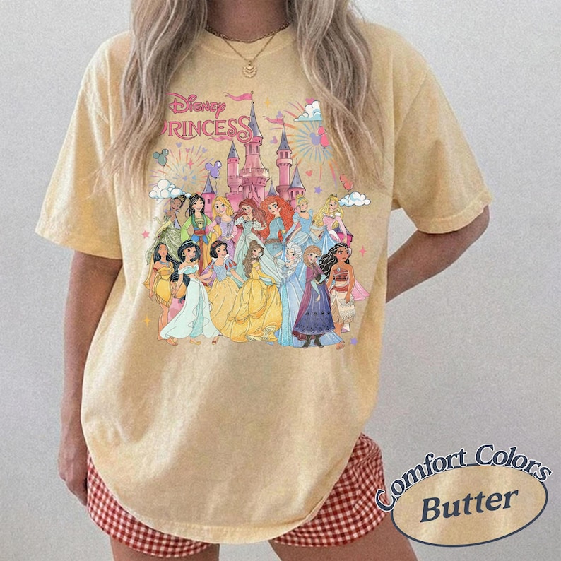May include: Butter-colored t-shirt featuring a graphic of Disney Princesses in front of a castle. The text "Disney Princess" is above the castle. The shirt is a Comfort Colors brand.