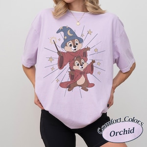 Op de afbeelding: Een lichtpaars T-shirt met een cartoonillustratie van twee chipmunks in tovenaarskostuums, een met een blauwe hoed en toverstaf. Op het shirt staan de woorden "Comfort Colors" en "Orchid".