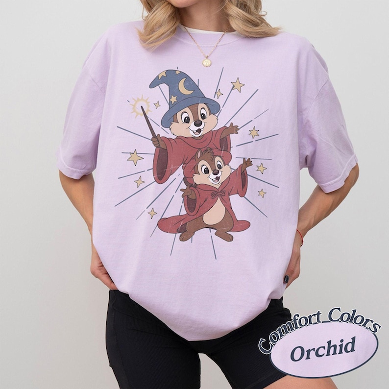 Op de afbeelding: Een lichtpaars T-shirt met een cartoonillustratie van twee chipmunks in tovenaarskostuums, een met een blauwe hoed en toverstaf. Op het shirt staan de woorden "Comfort Colors" en "Orchid".