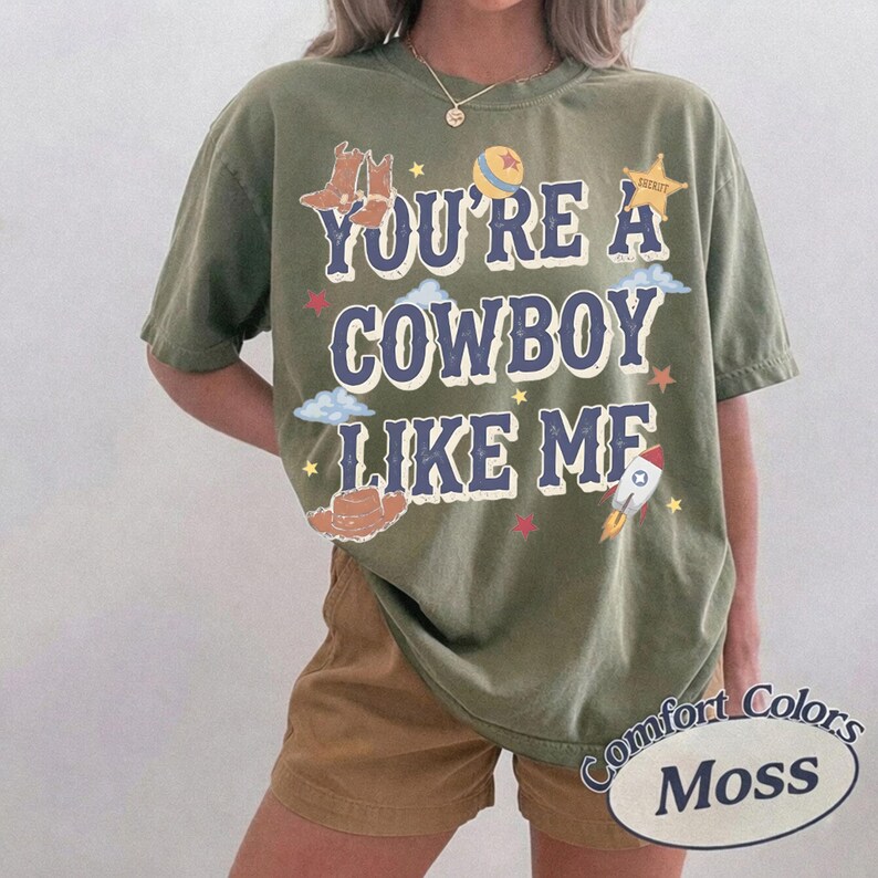 Op de afbeelding: Een olijfgroen T-shirt met korte mouwen en de tekst YOU'RE A COWBOY LIKE ME in donkerblauwe letters. Het ontwerp bevat illustraties van cowboylaarzen, een sheriffster, een bal, een cowboyhoed, wolken, sterren en een raket.
