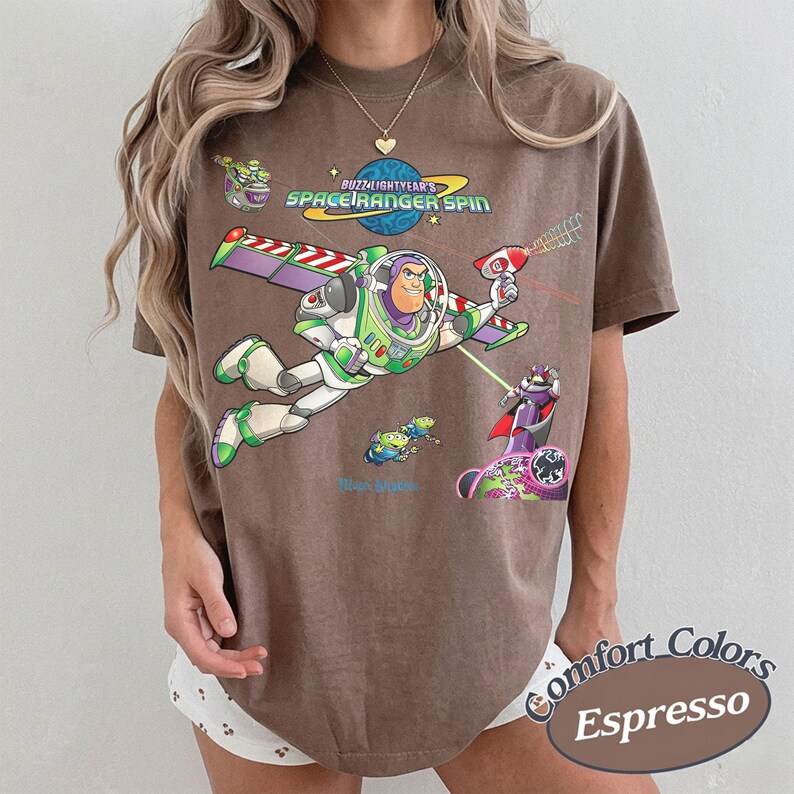 May include: Espresso-colored t-shirt featuring a graphic of Buzz Lightyear from Toy Story, with the text "Buzz Lightyear's Space Ranger Spin." The design includes various characters and elements from the movie. The shirt is part of the Comfort Colors collection.