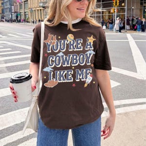 Op de afbeelding: Een donkerbruin T-shirt met korte mouwen en de tekst "YOU'RE A COWBOY LIKE ME" in een lichtblauw lettertype. Het ontwerp bevat illustraties van cowboylaarzen, een sheriffster, een raket, een bal en kleine gele, rode en witte sterren.