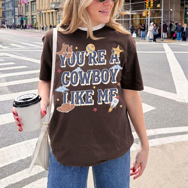 Op de afbeelding: Een donkerbruin T-shirt met korte mouwen en de tekst "YOU'RE A COWBOY LIKE ME" in een lichtblauw lettertype. Het ontwerp bevat illustraties van cowboylaarzen, een sheriffster, een raket, een bal en kleine gele, rode en witte sterren.