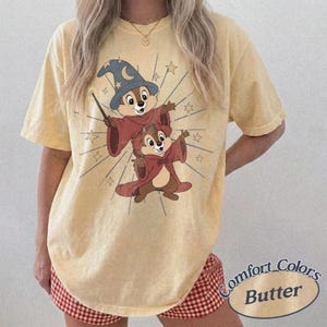Op de afbeelding: Lichtgeel T-shirt met een cartoonillustratie van twee chipmunks in tovenaarsgewaden, met een toverstaf en omringd door sterren. De woorden "Comfort Colors Butter" staan rechtsonder.