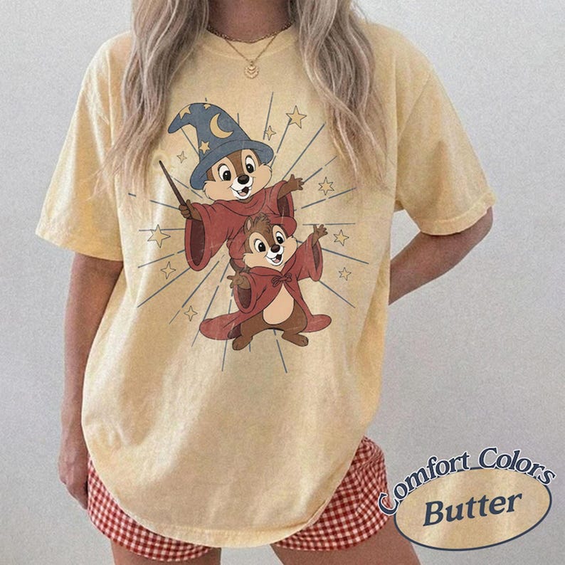 Op de afbeelding: Lichtgeel T-shirt met een cartoonillustratie van twee chipmunks in tovenaarsgewaden, met een toverstaf en omringd door sterren. De woorden "Comfort Colors Butter" staan rechtsonder.
