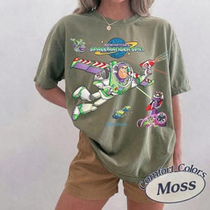 May include: Olive green t-shirt featuring a graphic of Buzz Lightyear from Toy Story, with the text "Buzz Lightyear's Space Ranger Spin." The design includes other characters and the Comfort Colors Moss logo.