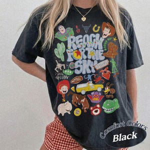 Peut inclure: Un t-shirt noir &agrave; manches courtes avec une impression graphique color&eacute;e portant le texte "REACH FOR THE SKY" entour&eacute; de diverses illustrations de personnages de dessins anim&eacute;s, dont un ranger de l'espace, un cow-boy, un camion jaune et un dinosaure vert.