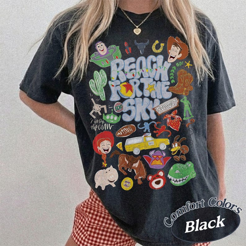 Peut inclure: Un t-shirt noir &agrave; manches courtes avec une impression graphique color&eacute;e portant le texte "REACH FOR THE SKY" entour&eacute; de diverses illustrations de personnages de dessins anim&eacute;s, dont un ranger de l'espace, un cow-boy, un camion jaune et un dinosaure vert.