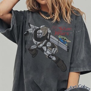 Pu&ograve; includere: T-shirt grigio scuro con una grafica di The Mandalorian e Grogu di Star Wars. Il design include il logo di Star Wars e il testo "Comfort Colors Pepper". La maglietta ha maniche corte e una vestibilit&agrave; ampia.