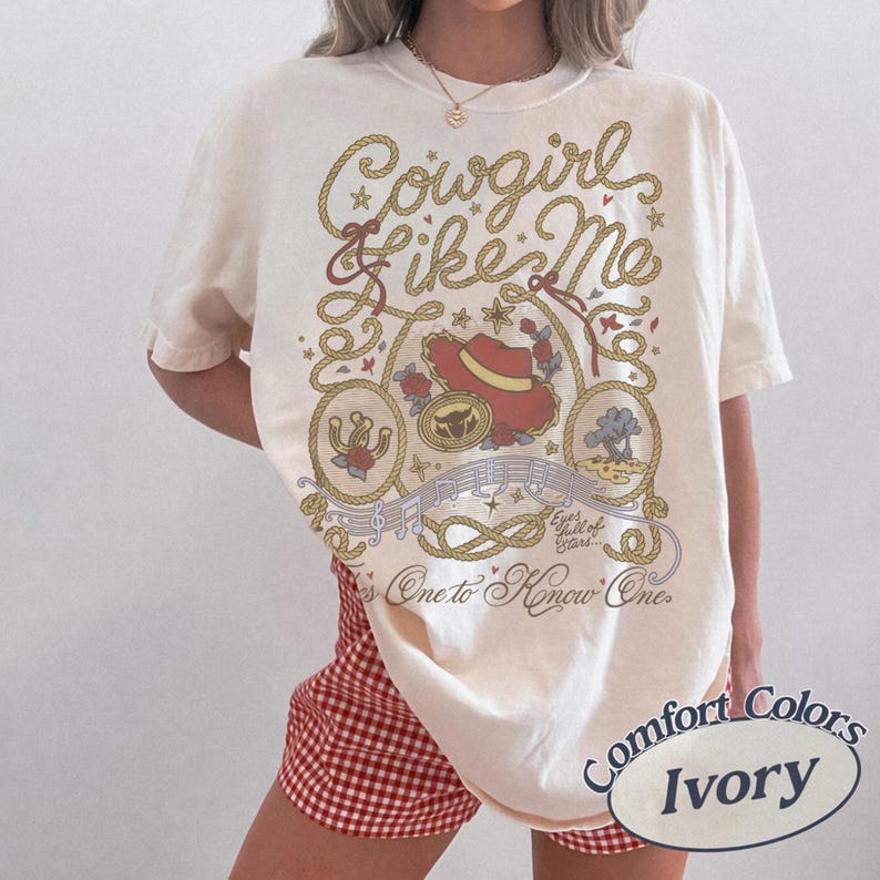 Op de afbeelding: Een ivoorkleurig t-shirt met een grafische print met de tekst Cowgirl Like Me, Eyes full of Stars en It's One to Know One. Het ontwerp bevat een rode cowboyhoed, een hoefijzer, een boom, muzieknoten en touwmotieven in bruin en rood.