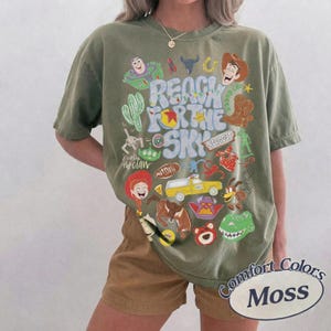 Peut inclure: Un t-shirt &agrave; manches courtes vert olive avec une impression graphique color&eacute;e repr&eacute;sentant divers personnages et ic&ocirc;nes de Toy Story. Le texte central indique Reach For The Sky dans une police bleue ondul&eacute;e, entour&eacute; de Woody, Buzz et Rex.