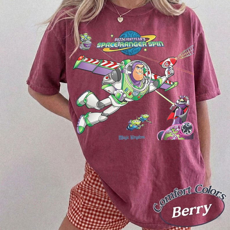 May include: Berry-colored t-shirt featuring a graphic of Buzz Lightyear in flight, with the text "Buzz Lightyear's Space Ranger Spin" and "Magic Kingdom." The design includes aliens and Zurg. The shirt is part of the Comfort Colors collection.