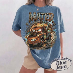 May include: A blue short-sleeve t-shirt featuring a graphic print of the character Mater from the movie Cars. The design includes the text MATER in a metallic font, surrounded by mechanical parts, gears, and blue lightning bolts on a dusty background.