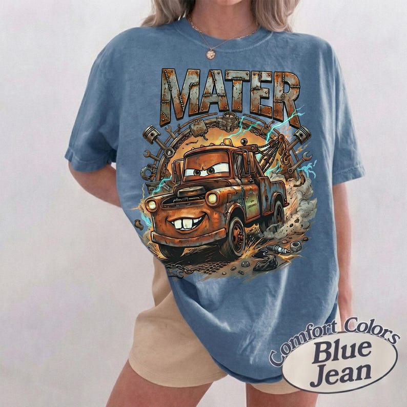 May include: A blue short-sleeve t-shirt featuring a graphic print of the character Mater from the movie Cars. The design includes the text MATER in a metallic font, surrounded by mechanical parts, gears, and blue lightning bolts on a dusty background.