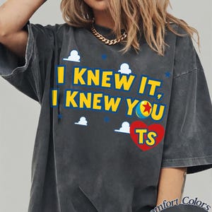 K&ouml;nnte beinhalten: Ein anthrazitfarbenes Kurzarm-T-Shirt mit dem gelben Text I KNEW IT, I KNEW YOU und einem roten Herz mit den Buchstaben TS. Das Design enth&auml;lt kleine wei&szlig;e Wolkensymbole und blaue Sterne sowie ein sichtbares Comfort Colors Pepper-Etikett.