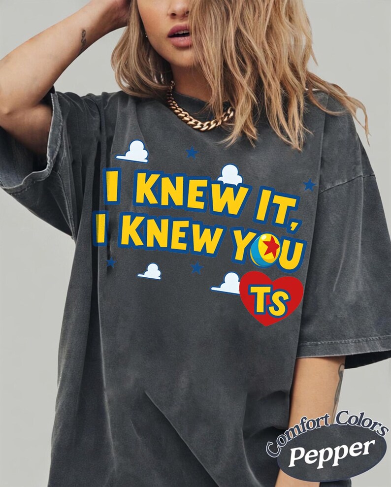 K&ouml;nnte beinhalten: Ein anthrazitfarbenes Kurzarm-T-Shirt mit dem gelben Text I KNEW IT, I KNEW YOU und einem roten Herz mit den Buchstaben TS. Das Design enth&auml;lt kleine wei&szlig;e Wolkensymbole und blaue Sterne sowie ein sichtbares Comfort Colors Pepper-Etikett.