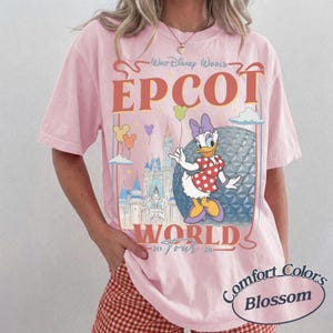 以下が含まれることがあります： Walt Disney World EPCOT World Tour 2026というテキストがプリントされたライトピンクのTシャツ。デイジーダック、エプコットのジオデシック・スフィア、城、ミッキー型の小さな風船のイラストが描かれています。