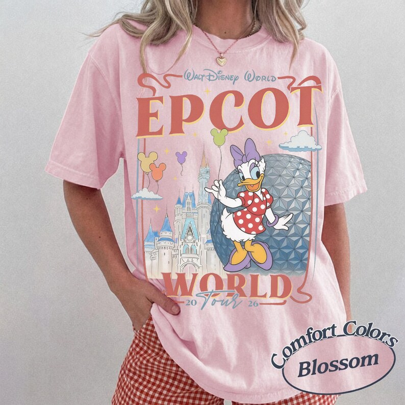 以下が含まれることがあります： Walt Disney World EPCOT World Tour 2026というテキストがプリントされたライトピンクのTシャツ。デイジーダック、エプコットのジオデシック・スフィア、城、ミッキー型の小さな風船のイラストが描かれています。