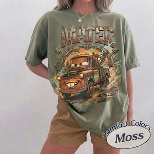 May include: An olive green short-sleeve t-shirt featuring a graphic print of a rusty tow truck character with the text MATER in large block letters above it. The design includes mechanical parts like gears and pistons in a circular arrangement.