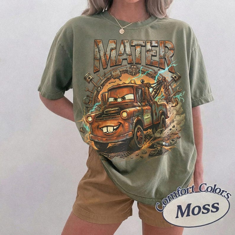 May include: An olive green short-sleeve t-shirt featuring a graphic print of a rusty tow truck character with the text MATER in large block letters above it. The design includes mechanical parts like gears and pistons in a circular arrangement.