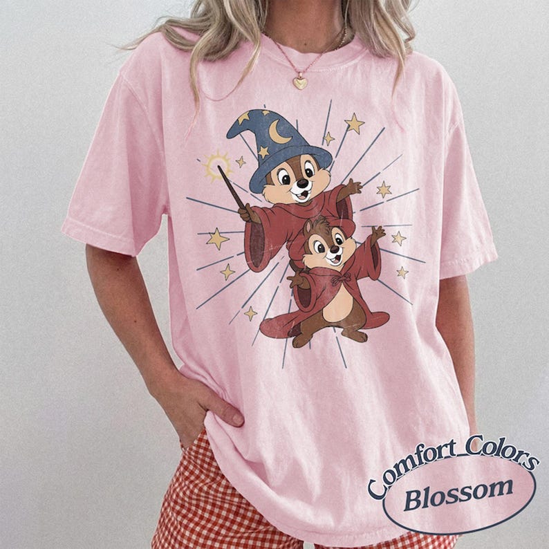 Op de afbeelding: Zachtroze Comfort Colors T-shirt met een cartoon illustratie van twee chipmunks verkleed als tovenaars, met een blauwe hoed en rode gewaden. Het ontwerp bevat sterren en een toverstaf. Het shirt heeft korte mouwen en een ronde hals.