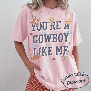 Op de afbeelding: Een lichtroze T-shirt met korte mouwen en de donkerblauwe tekst "YOU'RE A COWBOY LIKE ME" op de voorzijde. Het ontwerp bevat illustraties van bruine cowboylaarzen, een bal, een sheriffster, wolken, een hoed en een raket.
