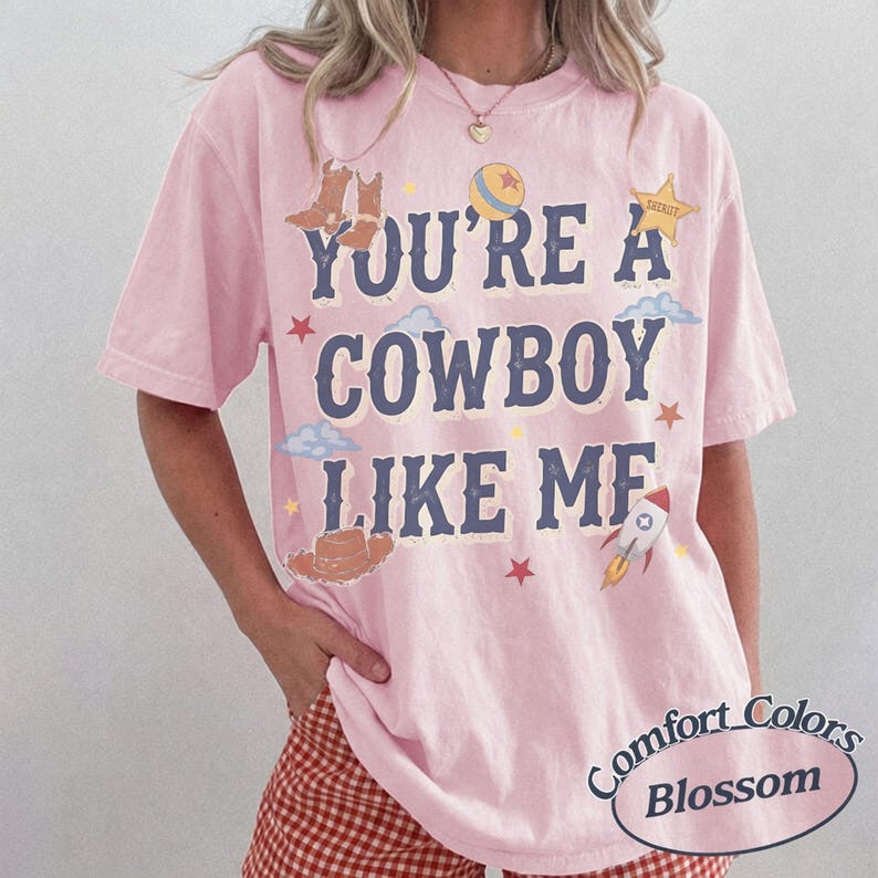 Op de afbeelding: Een lichtroze T-shirt met korte mouwen en de donkerblauwe tekst "YOU'RE A COWBOY LIKE ME" op de voorzijde. Het ontwerp bevat illustraties van bruine cowboylaarzen, een bal, een sheriffster, wolken, een hoed en een raket.