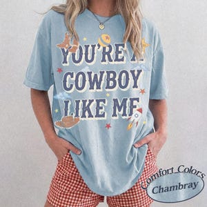Op de afbeelding: Een lichtblauw t-shirt met korte mouwen en de tekst "YOU'RE A COWBOY LIKE ME" in donkerblauwe letters. Het ontwerp bevat illustraties van een cowboylaars, een bal, een ster, een cowboyhoed en een raket in geel, rood en wit.