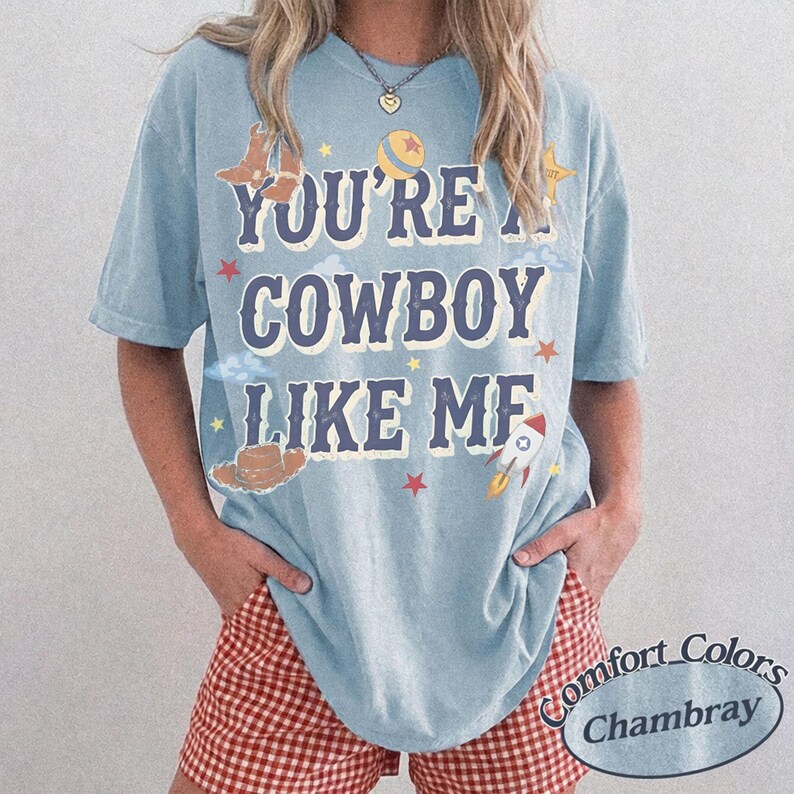 Op de afbeelding: Een lichtblauw t-shirt met korte mouwen en de tekst "YOU'RE A COWBOY LIKE ME" in donkerblauwe letters. Het ontwerp bevat illustraties van een cowboylaars, een bal, een ster, een cowboyhoed en een raket in geel, rood en wit.