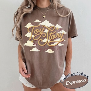 Może przedstawiać: Brązowy t-shirt z kr&oacute;tkim rękawem z grafiką białych chmur i napisem "Toy Story" wykonanym ż&oacute;łtą czcionką przypominającą linę. Poniżej gł&oacute;wnego tekstu znajduje się mniejszy napis kursywą "baby, just say yes". Koszulka ma okrągły dekolt.