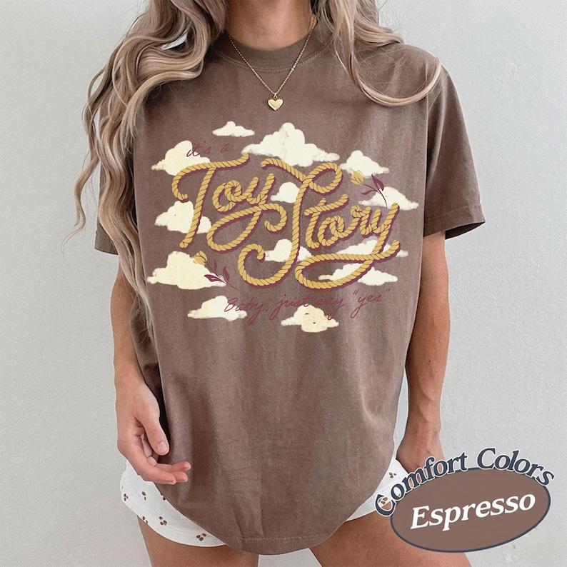 Może przedstawiać: Brązowy t-shirt z kr&oacute;tkim rękawem z grafiką białych chmur i napisem "Toy Story" wykonanym ż&oacute;łtą czcionką przypominającą linę. Poniżej gł&oacute;wnego tekstu znajduje się mniejszy napis kursywą "baby, just say yes". Koszulka ma okrągły dekolt.