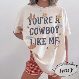 Op de afbeelding: Een ivoorkleurig t-shirt met de donkerblauwe tekst YOU'RE A COWBOY LIKE ME. De afbeelding bevat illustraties van bruine cowboylaarzen, een gele bal, een sheriffster, blauwe wolken, rode sterren, een bruine cowboyhoed en een raket.
