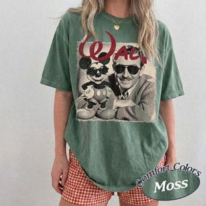 May include: A moss green t-shirt featuring a black and white image of Mickey Mouse and Walt Disney. The word "Walt" is printed in red script above the image. The shirt has a relaxed fit and is paired with red and white checkered shorts.