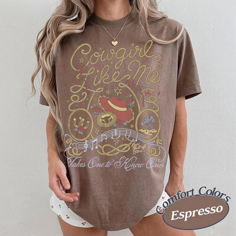 Op de afbeelding: Een bruin t-shirt met korte mouwen en een grafische print met de tekst Cowgirl Like Me en Takes One to Know One. Het ontwerp bevat een rode cowboyhoed, een lasso, muzieknoten en de zin Eyes full of Stars in wit en geel.