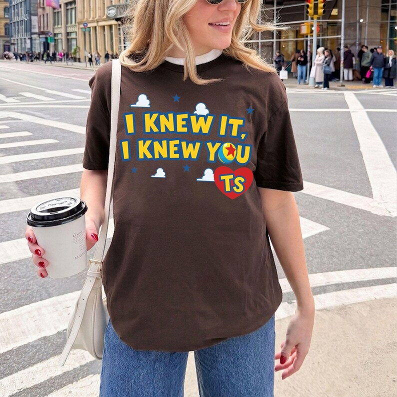 K&ouml;nnte beinhalten: Ein dunkelbraunes Kurzarm-T-Shirt mit dem gelben Schriftzug I KNEW IT, I KNEW YOU, einer bunten Ballgrafik und einem roten Herz mit den Buchstaben TS, umgeben von kleinen wei&szlig;en Wolken und blauen Sternen, die auf die Vorderseite gedruckt sind.