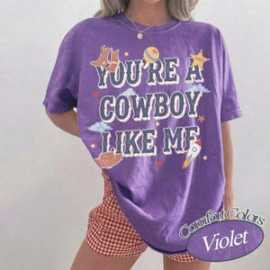 Op de afbeelding: Een paars t-shirt met korte mouwen en de tekst "YOU'RE A COWBOY LIKE ME" in een verweerd wit lettertype. De afbeelding bevat illustraties van cowboylaarzen, een bal, een sheriffster, wolken, een cowboyhoed en een raket.