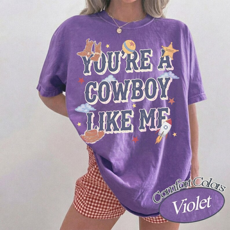 Op de afbeelding: Een paars t-shirt met korte mouwen en de tekst "YOU'RE A COWBOY LIKE ME" in een verweerd wit lettertype. De afbeelding bevat illustraties van cowboylaarzen, een bal, een sheriffster, wolken, een cowboyhoed en een raket.