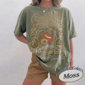 Op de afbeelding: Een olijfgroen t-shirt met korte mouwen en een grafische print met de tekst "Cowgirl like Me" en "One to Know One". Het ontwerp bevat een rode cowboyhoed, een lasso, muzieknoten en sterren in de kleuren geel, rood en wit op de stof.