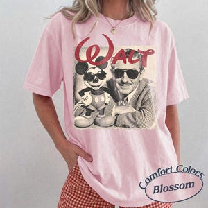 May include: Pink t-shirt featuring a vintage graphic of Mickey Mouse and Walt Disney with the word "Walt" in red. The shirt has a crew neck and short sleeves. The bottom right corner of the shirt has the words "Comfort Colors Blossom" in a circular design.