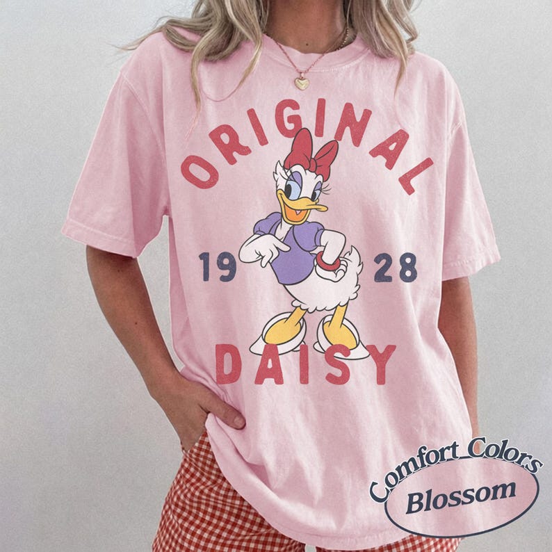 Op de afbeelding: Een lichtroze T-shirt met korte mouwen en een afbeelding van Daisy Duck met een rode strik, paarse top en gele schoenen. De tekst luidt ORIGINAL 1928 DAISY in rood en donkerblauw. Het shirt heeft een ronde hals en een losvallend silhouet.