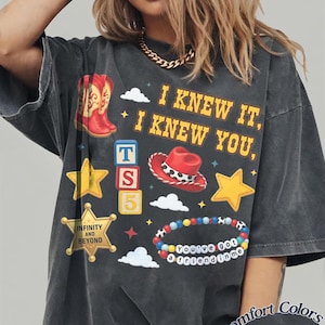 Pode incluir: Uma t-shirt cinzento-carv&atilde;o de manga curta com uma impress&atilde;o gr&aacute;fica com o texto I KNEW IT, I KNEW YOU, INFINITY AND BEYOND, e You've got a friend in me. O design inclui botas de cowboy vermelhas, um chap&eacute;u de cowboy vermelho, estrelas amarelas, blocos de letras e uma pulseira de contas colorida.