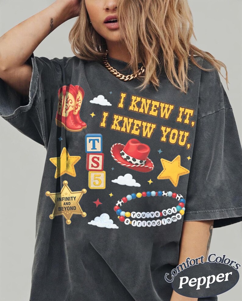 Pode incluir: Uma t-shirt cinzento-carv&atilde;o de manga curta com uma impress&atilde;o gr&aacute;fica com o texto I KNEW IT, I KNEW YOU, INFINITY AND BEYOND, e You've got a friend in me. O design inclui botas de cowboy vermelhas, um chap&eacute;u de cowboy vermelho, estrelas amarelas, blocos de letras e uma pulseira de contas colorida.