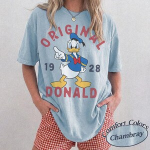 Op de afbeelding: Een lichtblauw T-shirt met korte mouwen en een afbeelding van Donald Duck die met zijn vinger wijst. De tekst ORIGINAL 19 28 DONALD staat in rode letters op het shirt. Het kledingstuk heeft een ronde hals en een relaxte pasvorm.