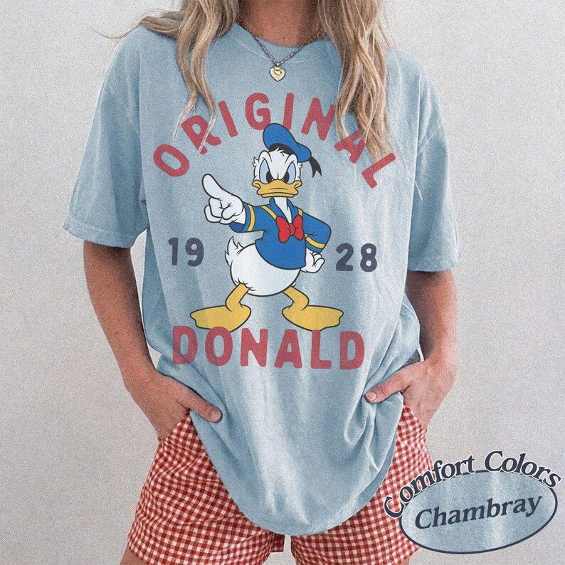 Op de afbeelding: Een lichtblauw T-shirt met korte mouwen en een afbeelding van Donald Duck die met zijn vinger wijst. De tekst ORIGINAL 19 28 DONALD staat in rode letters op het shirt. Het kledingstuk heeft een ronde hals en een relaxte pasvorm.