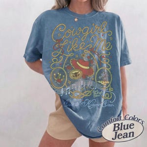 Op de afbeelding: Een blauw spijkerkleurig t-shirt met een grafische print van gele touwen, een rode cowboyhoed, muzieknoten en de tekst "Cowgirl Like Me", "Eyes full of stars" en "One to Know One". Het shirt heeft een ronde hals en korte mouwen.