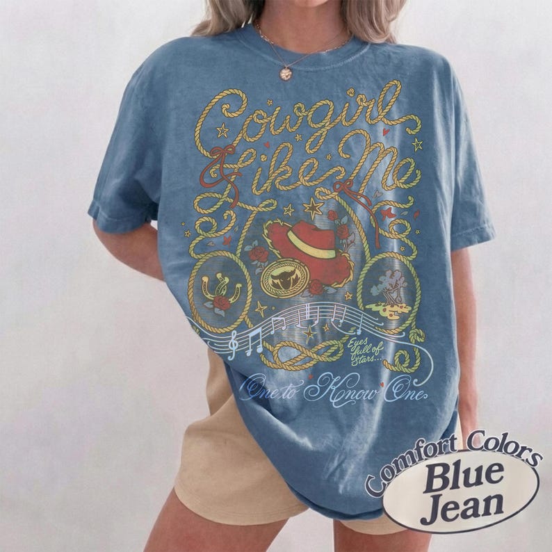 Op de afbeelding: Een blauw spijkerkleurig t-shirt met een grafische print van gele touwen, een rode cowboyhoed, muzieknoten en de tekst "Cowgirl Like Me", "Eyes full of stars" en "One to Know One". Het shirt heeft een ronde hals en korte mouwen.