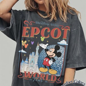 以下が含まれることがあります： Walt Disney World EPCOT World 20 Tour 26のテキストがプリントされたチャコールグレーの半袖Tシャツ。ミッキーマウス、エプコットのジオデシック・スフィア、城、そしてカラフルな風船のイラストが描かれています。